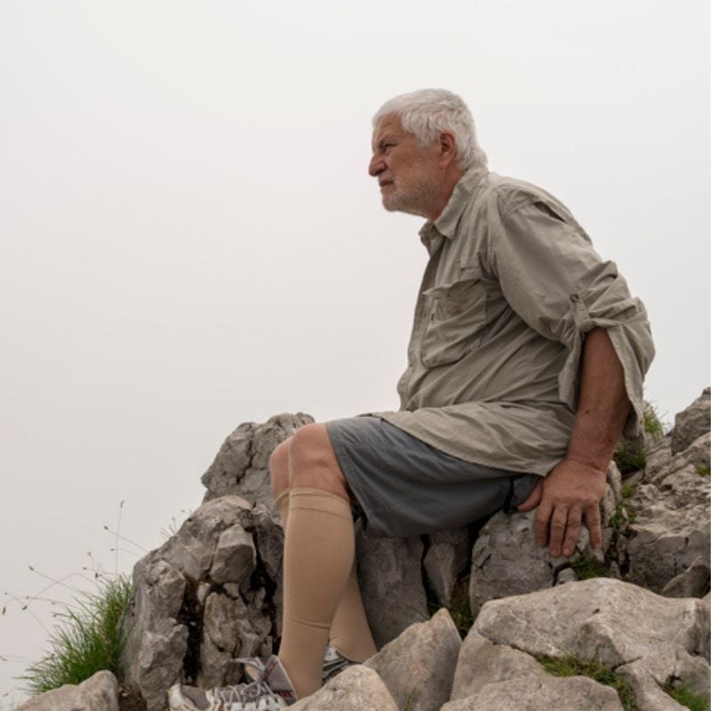 Older man sits on top of rocks during hike wearing hiking gear and tan compression stockings.