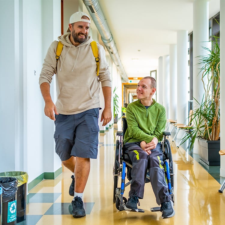 Caretaker walks beside patient in wheelchair moving together down hallway