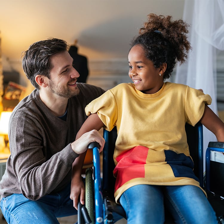 Man kneels beside young girl in wheelchair smiling and talking with her