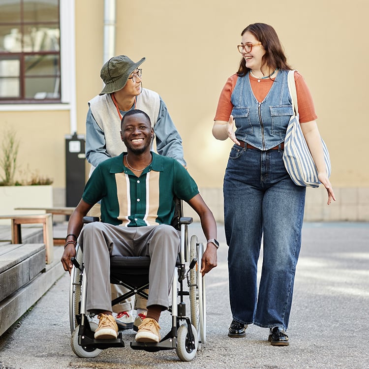Patient in wheelchair pushed by caregiver, walking alongside a smiling friend outdoors