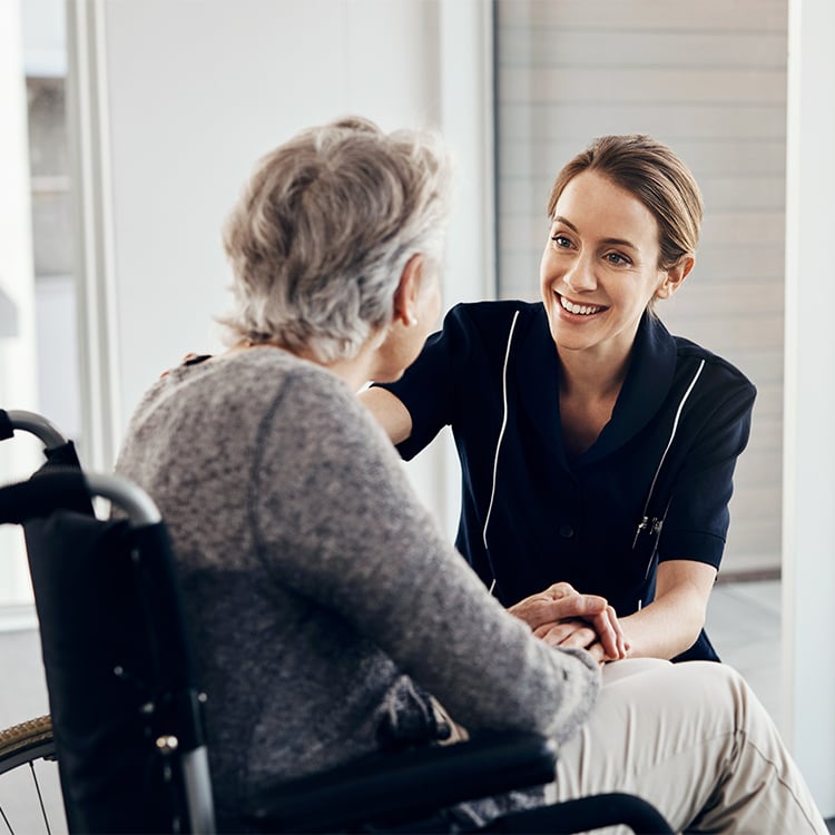 Caretaker kneels beside older woman in wheelchair smiling and speaking with her