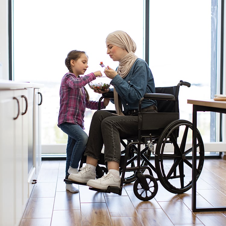 Woman in wheelchair sharing salad with young girl in kitchen
