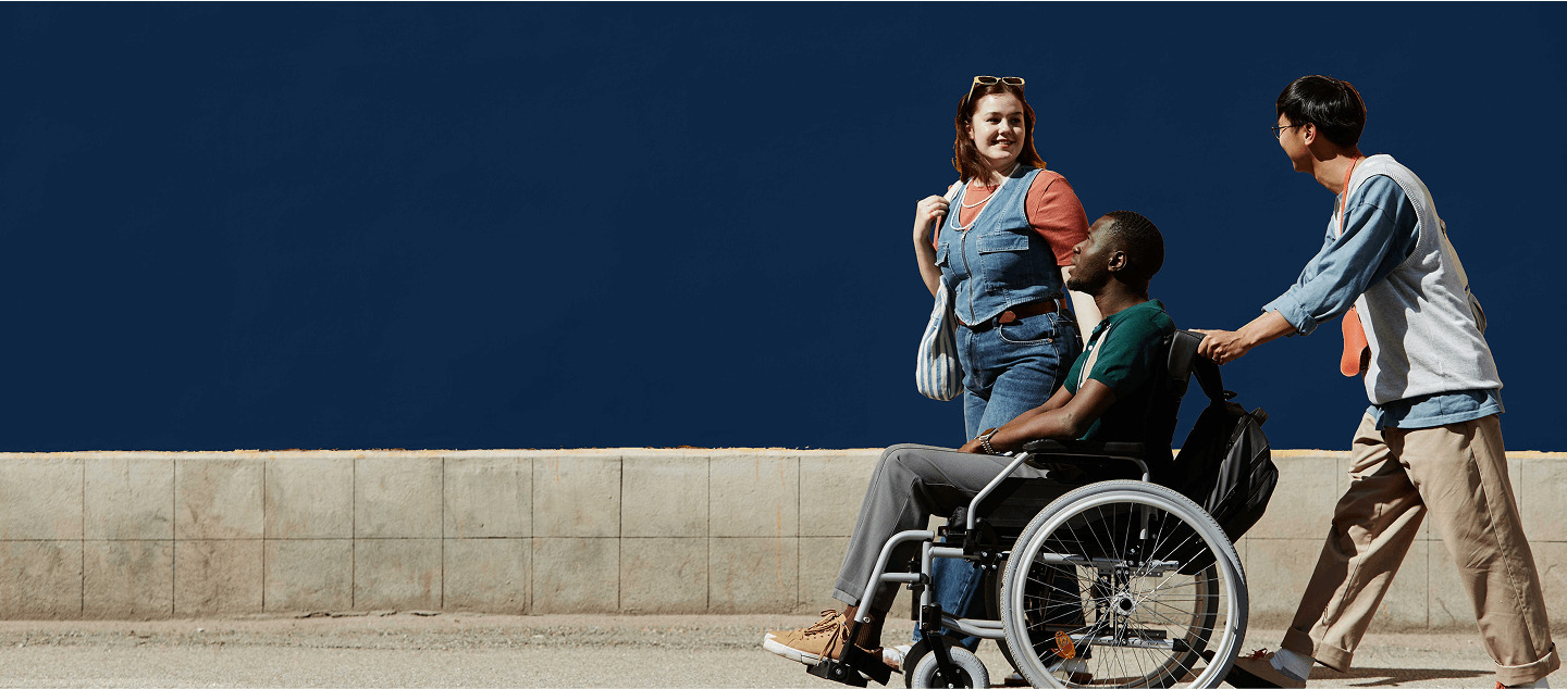 Patient in wheelchair pushed by caregiver, walking alongside a smiling friend