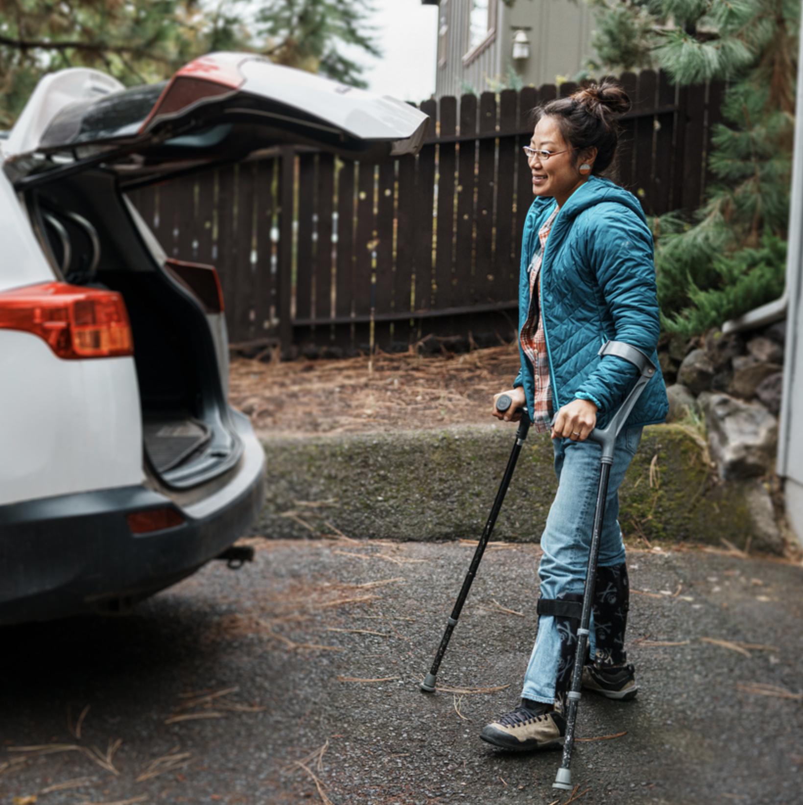 Woman walking with crutches approaches open trunk on car.