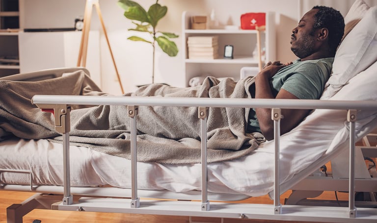 Older man sits in bed with blanket over his legs and guardrail beside him.