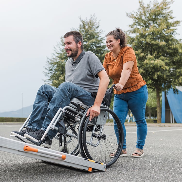 Caretaker pushes man in wheelchair up accessibility ramp with APA Medical support.=